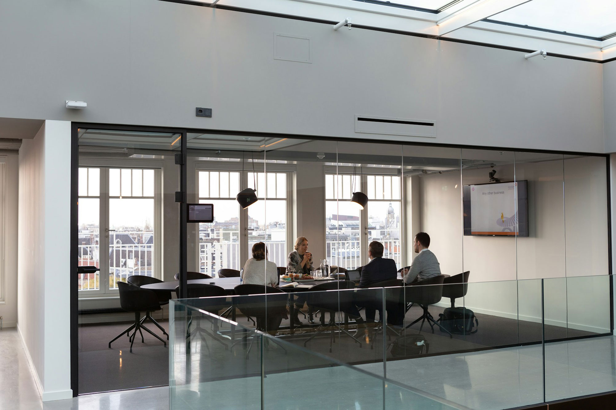 A modern office meeting room with glass walls. Inside the room, there are four people seated around a table, engaged in a discussion. The room has large windows that allow natural light to enter and provide a view of the outside. There is also a television screen mounted on one of the walls displaying some content.