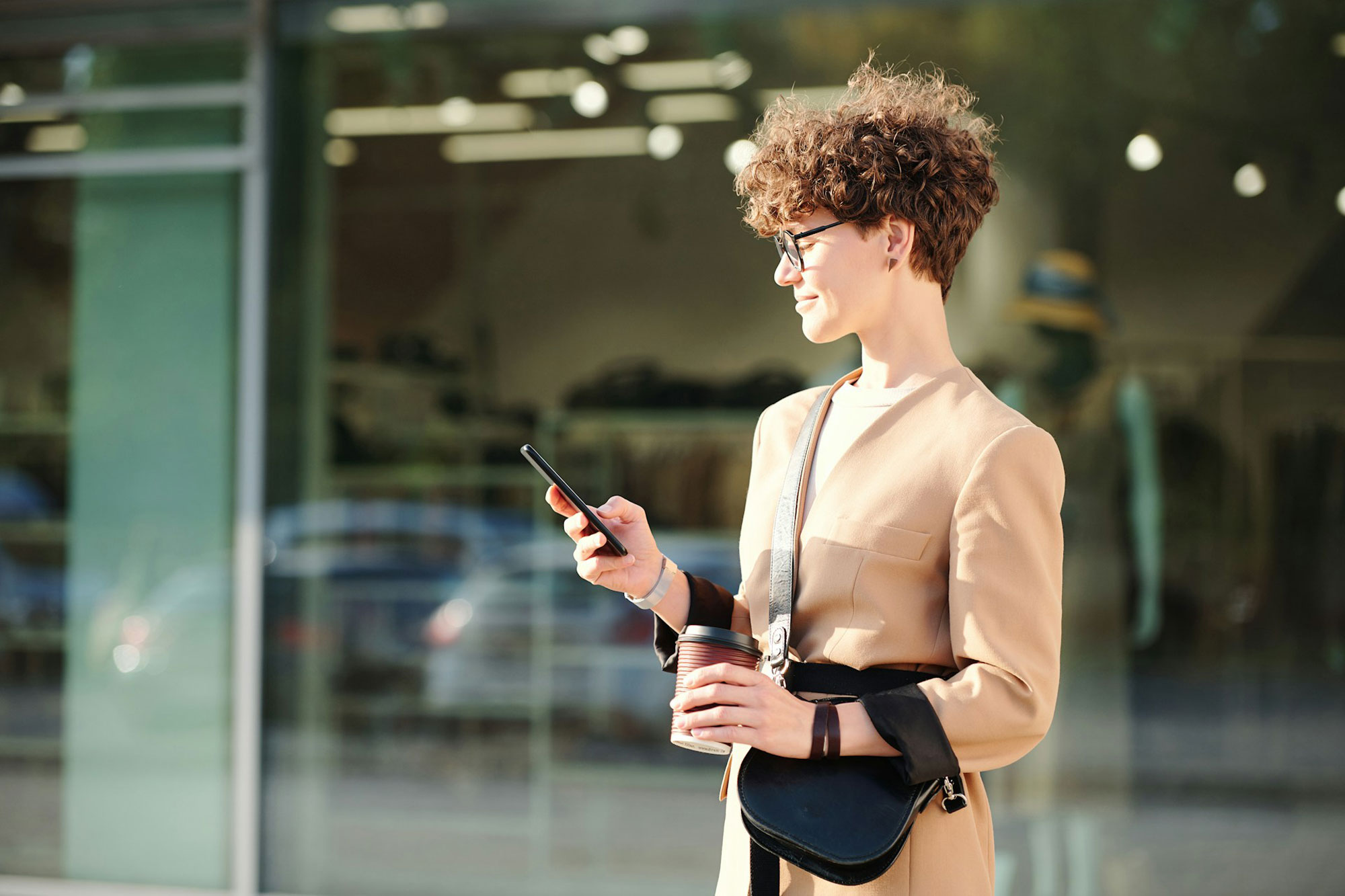 A person standing outdoors in front of a glass building, holding a smartphone in one hand and a coffee cup in the other. The person is wearing a beige coat and has a black crossbody bag. The person's face is pixelated for privacy