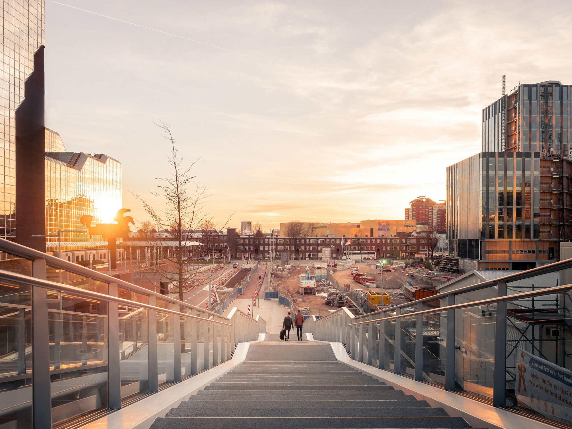 A modern urban scene at sunset, featuring a wide staircase leading down to a construction site with various buildings and cranes in the background. The sky is filled with warm colors from the setting sun, reflecting off the glass surfaces of nearby buildings. Two people are walking down the stairs towards the construction area.