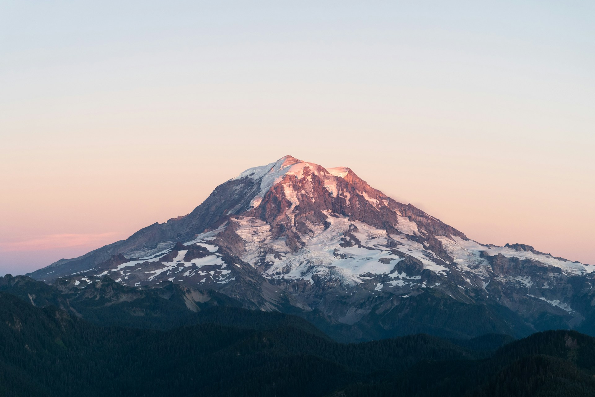 A snow-capped mountain with a pinkish hue at the peak, likely due to the setting or rising sun. The sky is clear with a gradient from light blue to soft pink. The foreground consists of dark, forested hills or mountains.